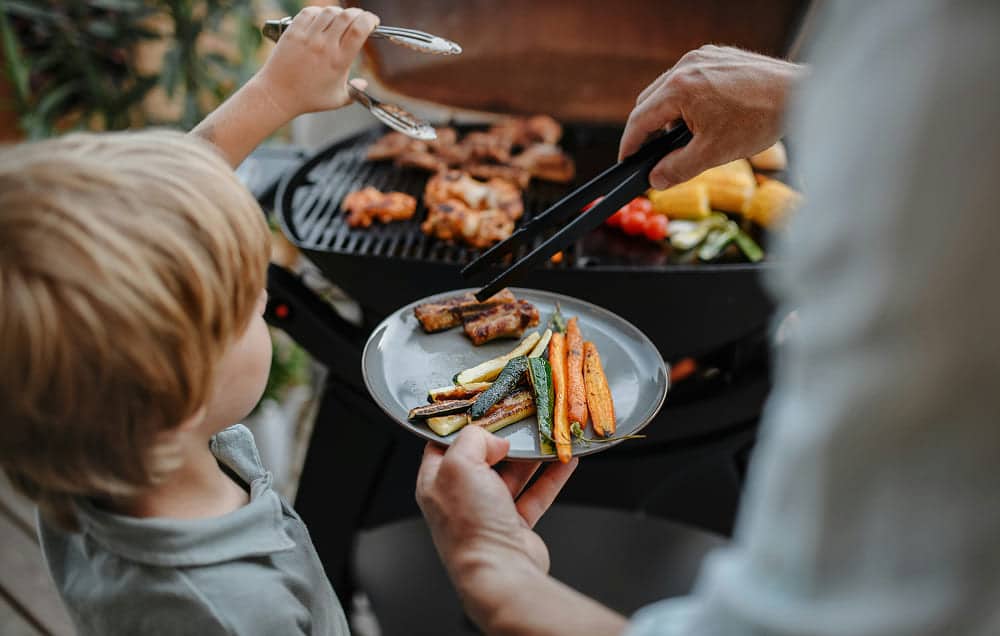 A child and adult barbequing together, viewed from behind.